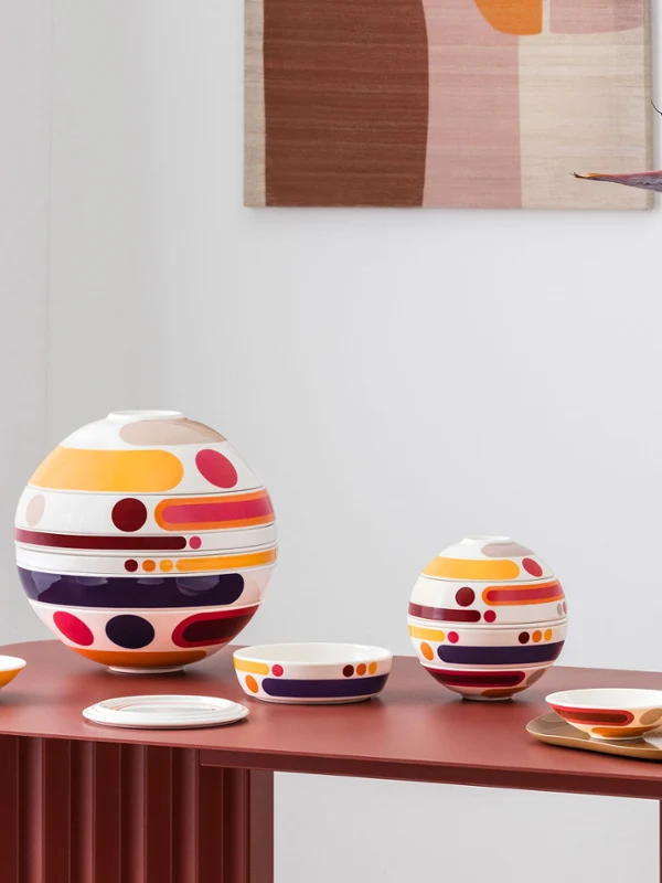 Des services de table colorés et composés de plusieurs pièces de la petite boule miami, objets design circulaires de grande et petite taille, sont disposés à titre décoratif à côté d'un vase aux motifs abstraits orné de fleurs orange sur une table rouge.