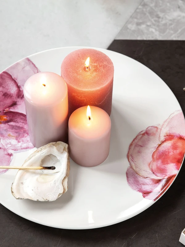 Une assiette décorative Rose Garden avec des bougies allumées à côté d’un coquillage et d’un livre avec un motif réalisé à la feuille d’or.