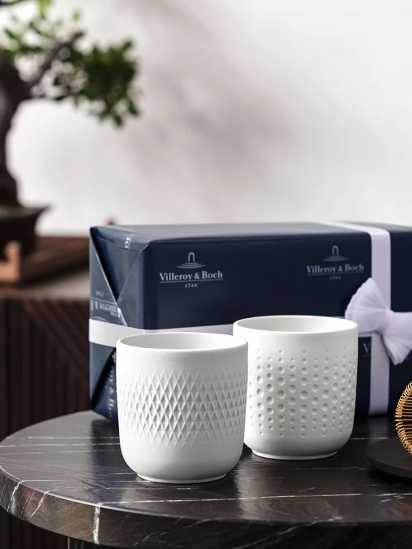 Two white collier cups in front of a bonsai tree Two white collier cups on a table with a tea whisk and a gift box, in front of a bonsai tree.