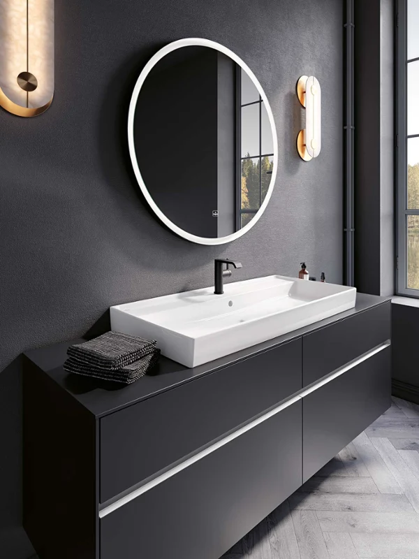 Une salle de bains moderne avec un élégant lavabo noir, une vasque blanche Collaro de Villeroy & Boch, un miroir rond rétroéclairé et des luminaires fixés au mur. Une grande fenêtre permet de bénéficier de la lumière naturelle et d’une vue sur l’extérieur.