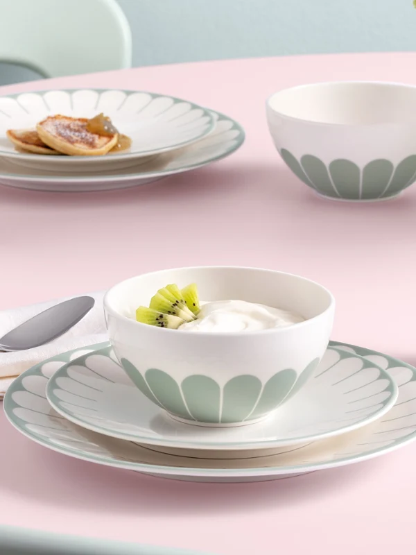 Un service de table composé d'assiettes, de bols et de tasses ornés d'un motif floral, avec de la nourriture et un vase en verre Fleur sur une nappe rose.