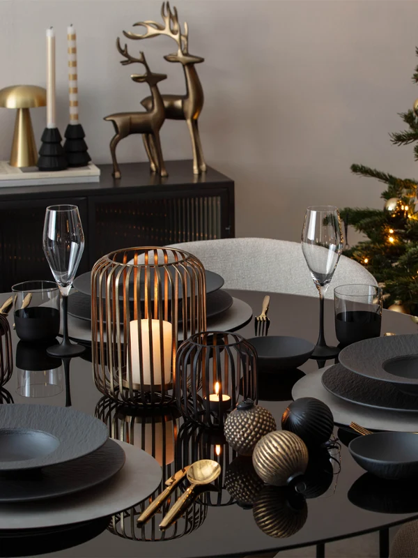 A black dining table set with dark crockery, Christmas decorations, candles and an illuminated Christmas tree in the background.