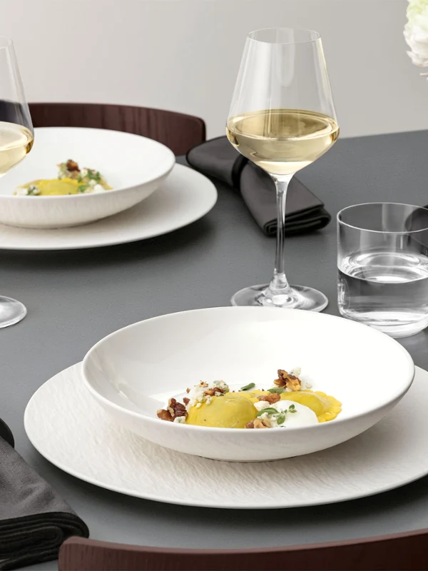 A formal table setting with a Manufacture bowl on top of a plate A formal table setting with a white bowl on a white plate, glassware, and silver cutlery on a grey table and food.