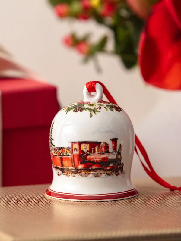Eine Keramikglocke mit einem Zugmotiv sitzt auf einer goldenen Schachtel, mit einem roten Geschenk und Blumen im Hintergrund.