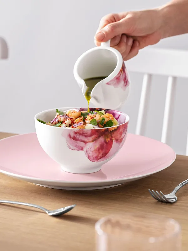Eine Person gießt Dressing auf einen Salat in einer Rose Garden Schüssel an einem elegant gedeckten Esstisch mit Rose Garden Geschirr.