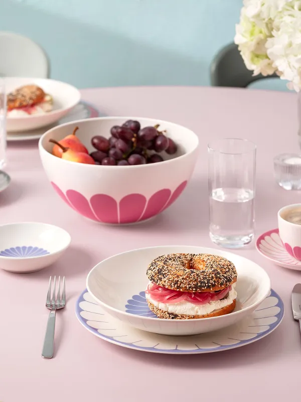 Ein runder rosa Tisch mit Tellern voller Essen, darunter ein belegter Bagel, eine Obstschale, Tassen Kaffee und Besteck. Im Hintergrund steht eine Vase mit weißen Blumen.