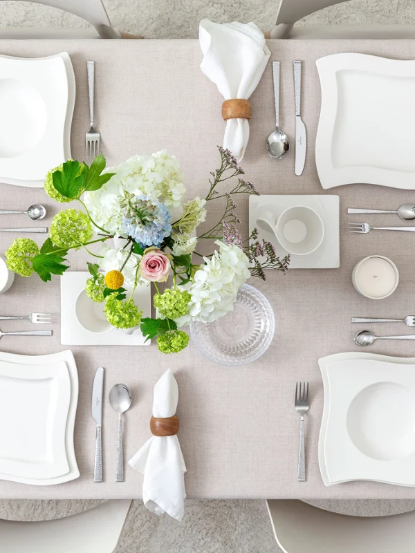 Draufsicht auf ein minimalistisches Tischgedeck mit NewWave Porzellantellern, Kaffeetasse und Untertasse, Boston- und Boston-Green-Gläsern sowie NewWave- und Blacksmith-Besteck. Blumenvasen setzen einen natürlichen und eleganten Akzent in der klaren Komposition.