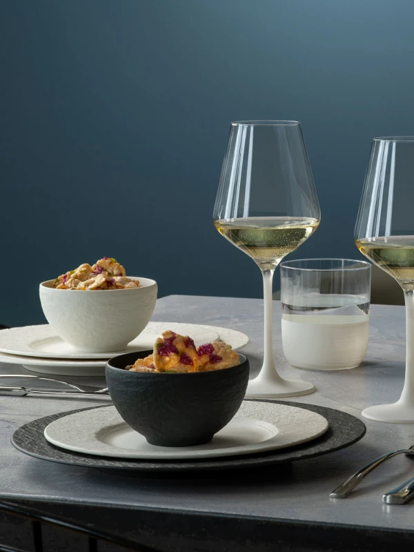 Un dîner élégant avec deux verres à vin blanc, des assiettes et des plats creux Manufacture disposés de façon ordonnés et contenant des mets sur une table moderne.