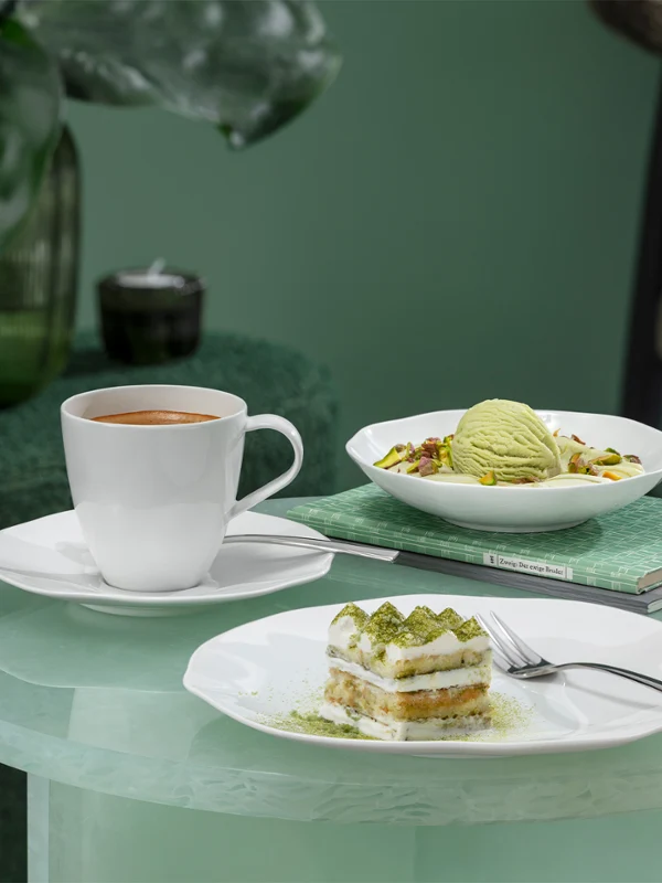 Une tasse à café Pura, une part de gâteau et une coupe à dessert Pura avec de la glace sont posées sur une table en verre avec une plante verte en arrière-plan.