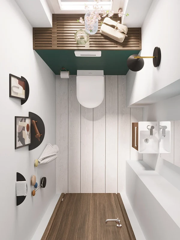 Vue de dessus d’une petite salle de bains avec lavabo mural, miroir, toilettes et décor moderne d’art abstrait. La pièce a un sol et des murs blancs et une porte en bois.