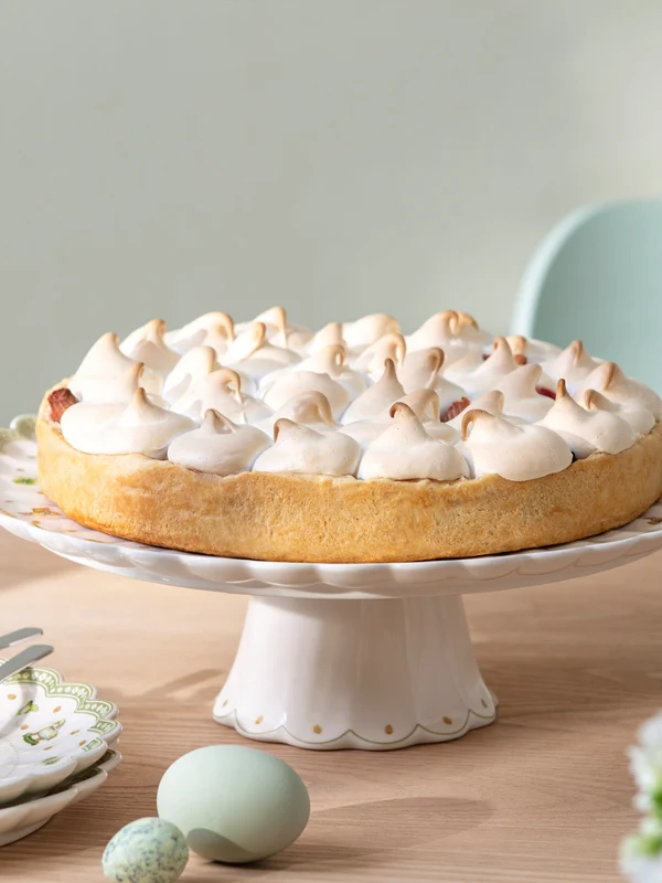 Auf einem Holztisch steht eine Torte mit Baiser auf einem Tortenständer mit Osterdekoren von Easter Delight, daneben stehen Teller und Blumen.