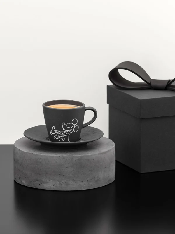 Black coffee cup and saucer on a grey pedestal Black coffee cup and saucer on a grey pedestal next to a black gift box with a bow on a black surface.