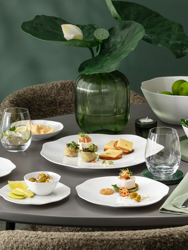 Une table ronde dressée avec la vaisselle blanche de la collection Pura, comprenant des assiettes à entrée, des verres, des serviettes et un centre de table composé de plantes vertes.