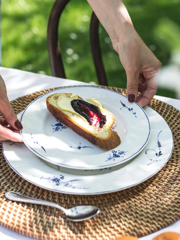 Eine Person stellt einen Old Luxembourg Teller, belegt mit Fruchtaufstrich und Butter, auf ein gewebtes Tischset.