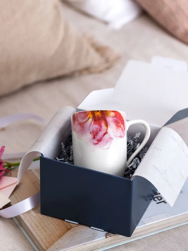 Eine offene Villeroy & Boch Geschenkbox mit einer Rose Garden Tasse, rosa Blumen und einer Notiz auf einem Holztablett, im Hintergrund weiche Kissen.