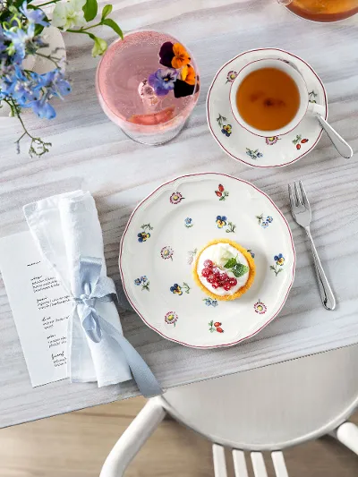 Petite Fleur Teller mit einer Torte, einer Teetasse, einem Getränk mit einer Blume und einer Serviette auf einem Holztisch.