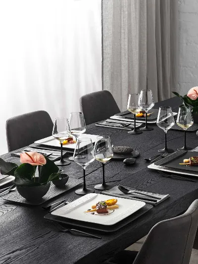 Ein schwarzer Tisch in einem eleganten Wohnzimmer gedeckt mit Gläser, rechteckigem Geschirr und Besteck aus der Kollektion Manufacture von Villeroy & Boch.