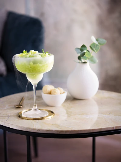 A chilled cocktail, garnished with lime twist, served in a Purismo cocktail glass on a marble table, accompanied by a small serving bowl of snacks and a white vase with green plants.