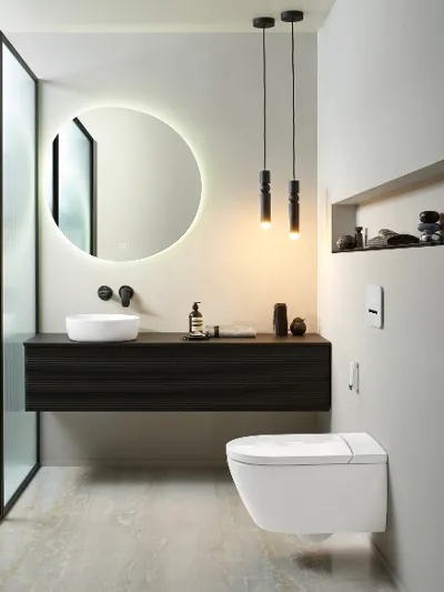 Antao Kollektionsbad mit ViClean Dusch-WC von Villeroy & Boch Modernes Badezimmer mit wandmontiertem WC, schwebendem schwarzen Waschtisch mit weißem Becken, beleuchtetem runden Spiegel, Hängelampen und Trennwand aus Milchglas.