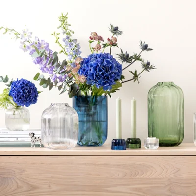 Un sideboard avec des vases Fleur de Villeroy & Boch, ornés de fleurs bleues et violettes, à côté de deux bougies vertes et de livres empilés.