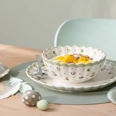 Un bol décoratif Easter Delight contenant des fruits et du yaourt est posé sur une assiette avec une cuillère, à côté d'une tasse et d'une soucoupe, sur une table.