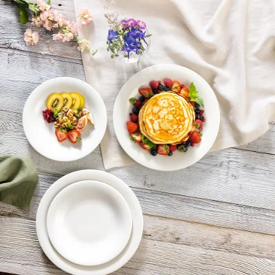 Una pila di pancake con frutti di bosco viene servita su un piatto New Cottage di Villeroy & Boch, con frutta a fette su un altro piatto. Il tavolo di legno è decorato con fiori e una tovaglia drappeggiata.