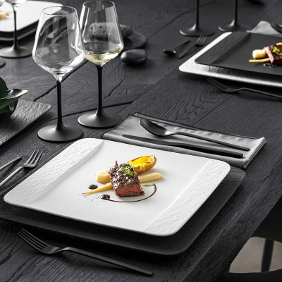 De la vaisselle Manufacture en noir et blanc avec un plat principal servi sur une table noire, à côté de verres à pied noirs de Villeroy & Boch.