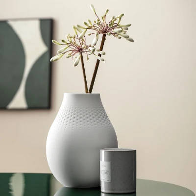 A white Manufacture vase with a subtle flower arrangement stands on a green table.