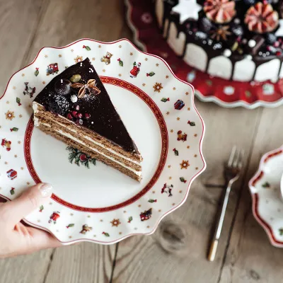 A hand holding a plate with a slice of chocolate cake. On the table there is also a decorated complete cake, a cup of coffee, a fork and a saucer and plate set from Toy's Delight.