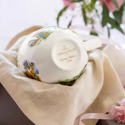 Une tasse de l'Annual Easter Edition de Villeroy & Boch est posée à l'envers sur un tissu beige et montre son cachet annuel doré, tandis qu'une fleur rose et un vase avec des feuilles sont visibles en arrière-plan.	