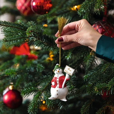 A hand hangs the Villeroyand Boch ornament, a snowman with a broom and a present, on a decorated Christmas tree adorned with red baubles and lights.