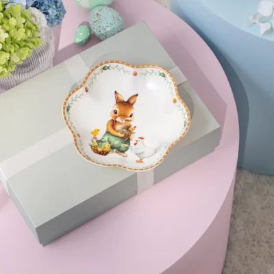 Une boîte cadeau avec un ruban posée sur une table rose, surmontée d’une assiette décorative Annual Easter Edition 2026 avec un lapin et des fleurs.