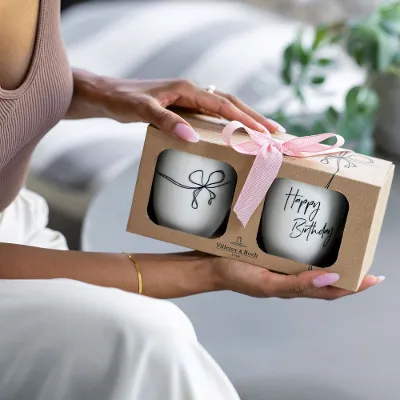 Person holding a gift box with a pink bow containing two 'Happy Birthday' Statement mugs.