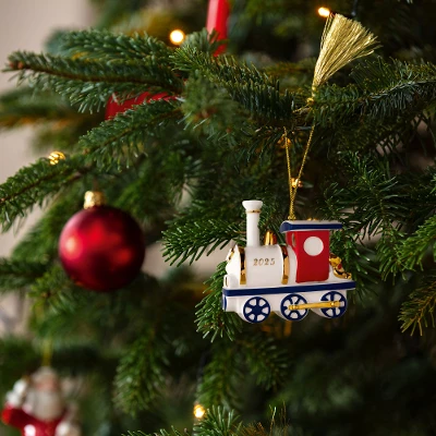 Christmas Classics 2025 Train ornament on tree  A Christmas tree adorned with a festive train ornament marked "2025" and red baubles. Warm lights add a cozy, cheerful holiday atmosphere.