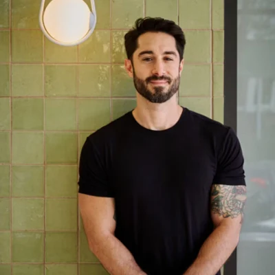 A bearded man in a black t-shirt stands before a green tiled wall with a white globe pendant light, wearing a watch on his left wrist and a tattoo.