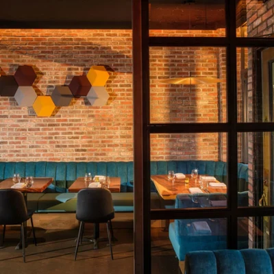 A restaurant interior with exposed brick walls, teal tufted booths, wooden tables, black chairs, and geometric wall art under warm ambient lighting.