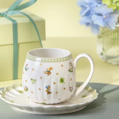Tasse à thé Easter Delight sur une soucoupe Easter Delight avec un coffret cadeau et un vase de fleurs bleues en arrière-plan.