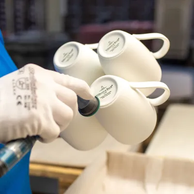 Cups with footstamp in one hand with a white glove. A hand with a white glove uses a tool to polish the foot of the 4 cups; the Villeroy & Boch stamps are visible on the bottom of the cups.