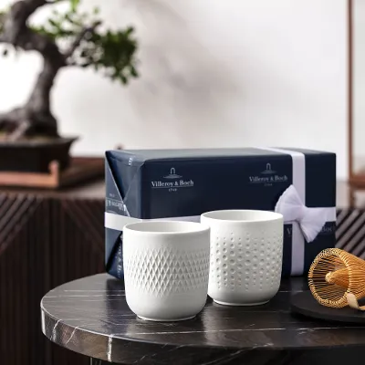 Two white collier cups on a table with a tea whisk and a gift box, in front of a bonsai tree.