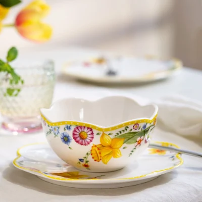 La tasse à thé et la soucoupe « Spring Awakening » avec cuillère de Villeroy & Boch sont posées sur la table, à côté de verres en cristal et de fleurs doucement floues en arrière-plan.