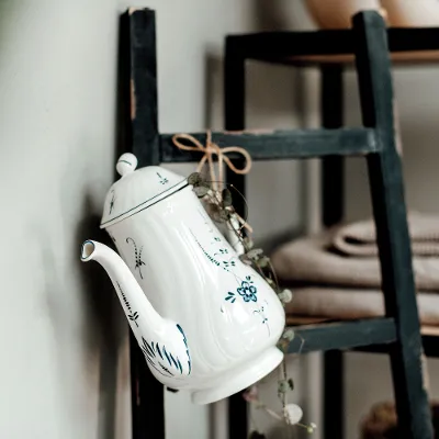 Een witte en blauwe Vieux Luxembourg theepot met bloemenmotief hangt naast opgevouwen handdoeken aan een zwarte houten ladder.