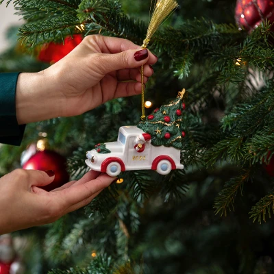 A person hangs a small ornament in the shape of a Villeroy & Boch truck on a decorated Christmas tree adorned with red baubles and lights.