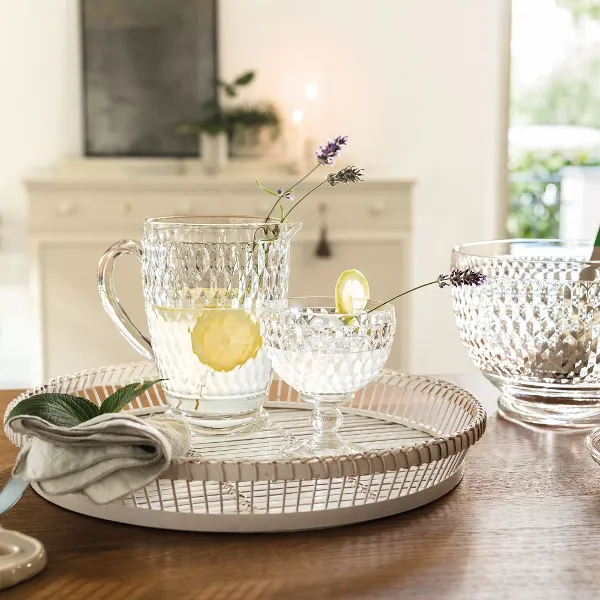 Boston-clear-jug-champagne-salad bowl Glasses from Villeroy & Boch with lemon water stand on a tray on a wooden table with glass bowls and a napkin.