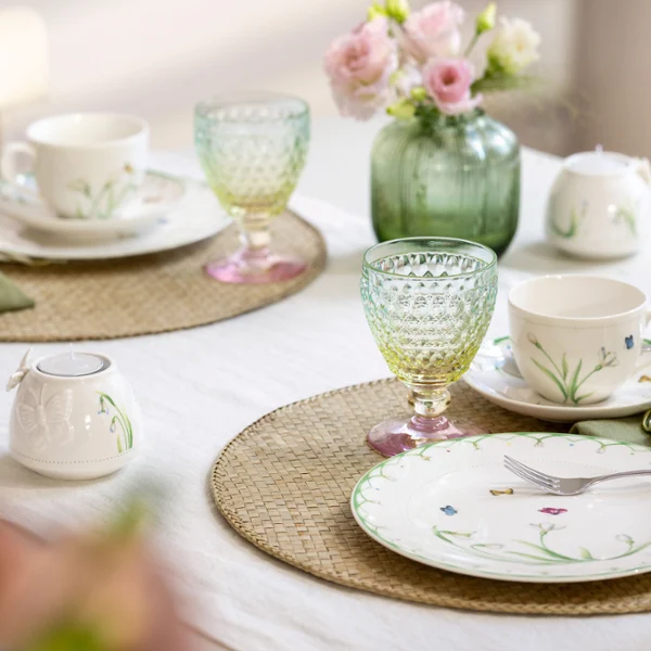 The Colourful Spring set from Villeroy & Boch embellishes any table for two with plates and cups with a floral pattern, bright green and yellow glasses, pink flowers in a vase and beige placemats on a white tablecloth.