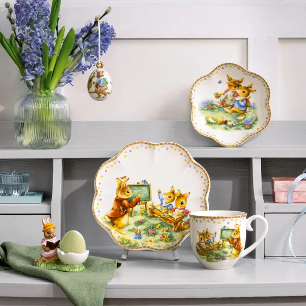 De Annual Easter Edition van Villeroy & Boch presenteert decoratieve borden en een mok met dierenmotieven op een commode. Een kleine eierdop met een beeldje en een vaas met blauwe bloemen maken het arrangement compleet.	