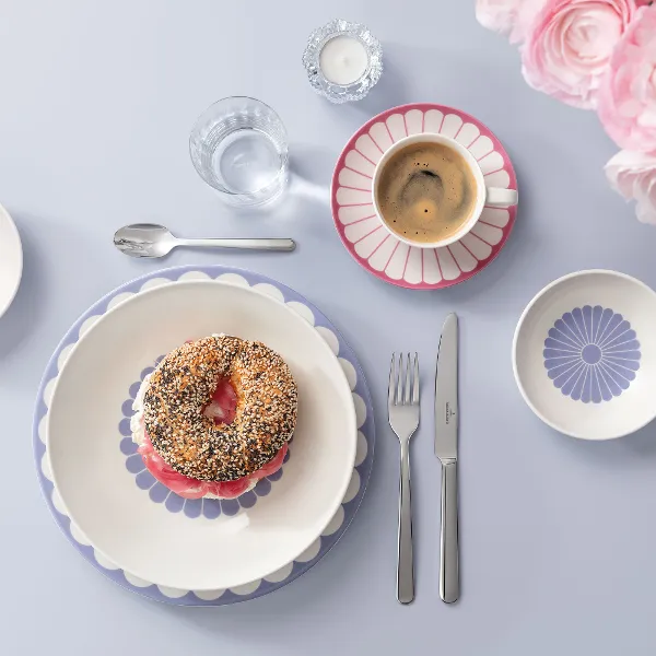 Ein Frühstücksgedeck mit Bagel, Tasse Kaffee, dekorativen Tellern, Besteck und Blumen auf hellblauem Untergrund. Ein Frühstücksgedeck mit Bagel, Tasse Kaffee, dekorativen Tellern, Besteck und Blumen auf hellblauem Untergrund.
