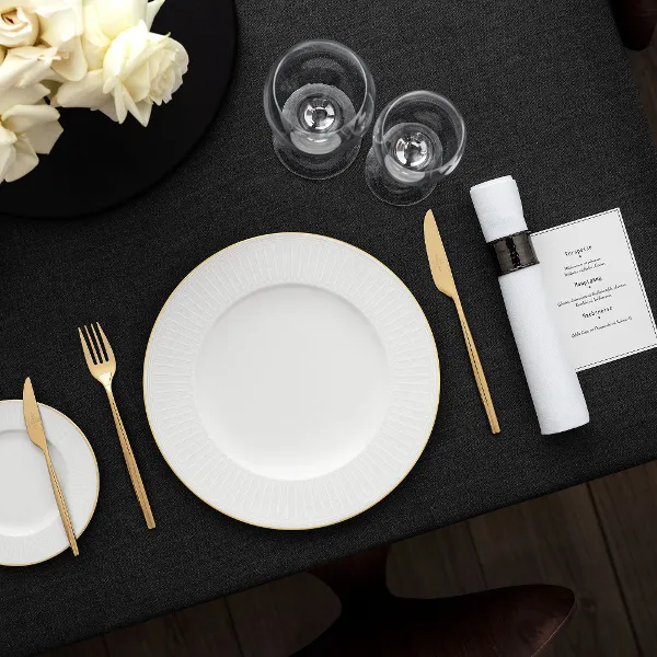 Une table dressée avec des assiettes blanches Château Septfontaines, des couverts de table dorés, deux verres, une serviette roulée et une composition florale sur une nappe foncée.