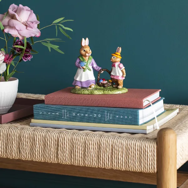 Two decorative bunny figurines from the Villeroy & Boch Bunny Tales collection on stacked books on a woven bench, next to a vase with buttercups and roses.