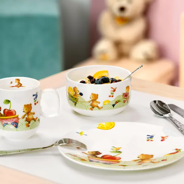 Un ensemble pour le petit-déjeuner Hungry as a bear avec un bol de céréales et des fruits sur une table, ainsi qu’une tasse, des couverts de table et un ours en peluche en arrière-plan.