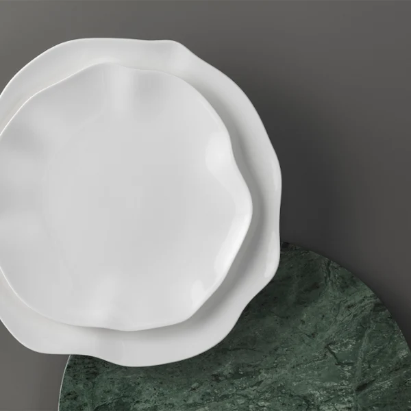 Deux assiettes Pura blanches de forme irrégulière sont empilées sur une assiette ronde vert foncé devant un fond gris.